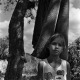 Black and white portrait photo of young girl standing before drying old carpet in backyard with a boy hiding behind tree