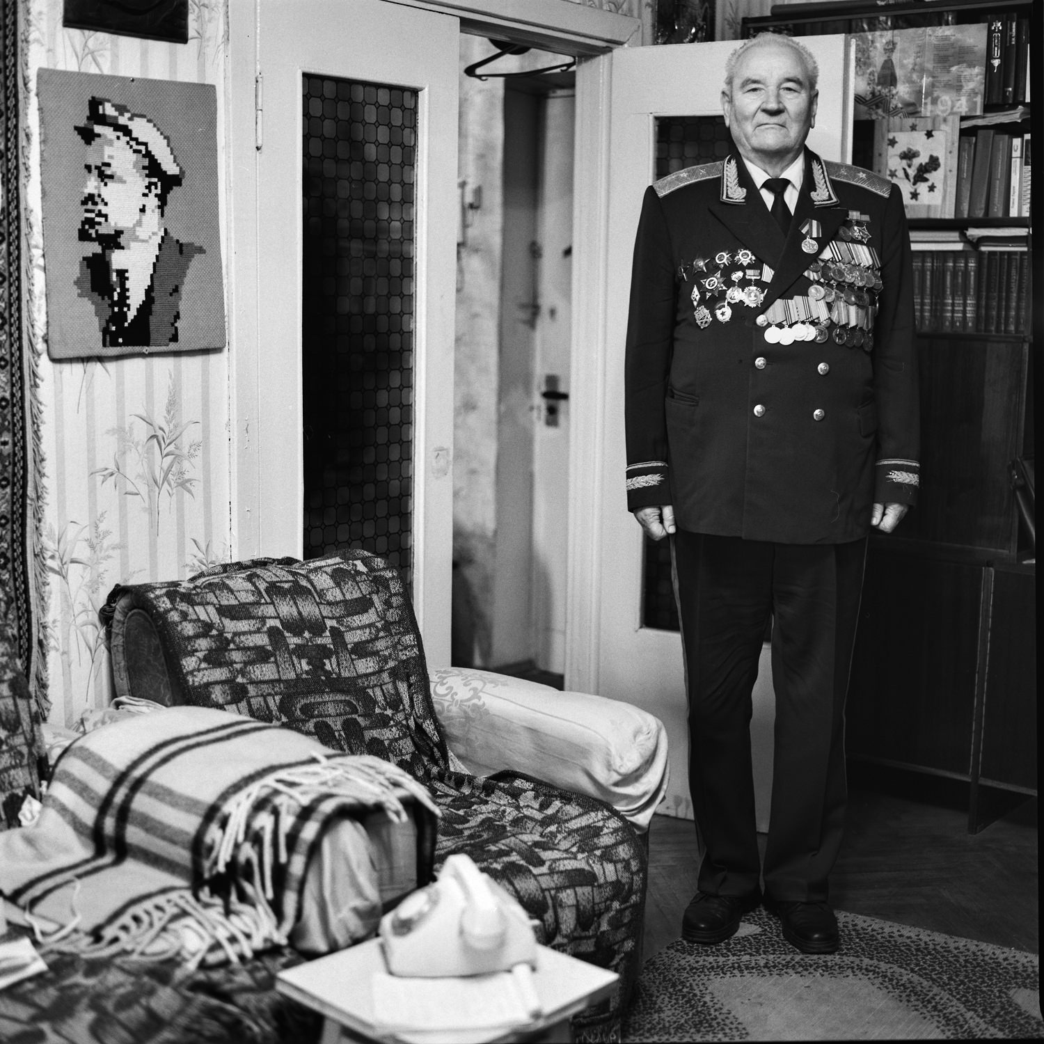 Portrait of the Great Patriotic War veteran in unifrm with medals standing still in his room with hand-made picture of Lenin on wall