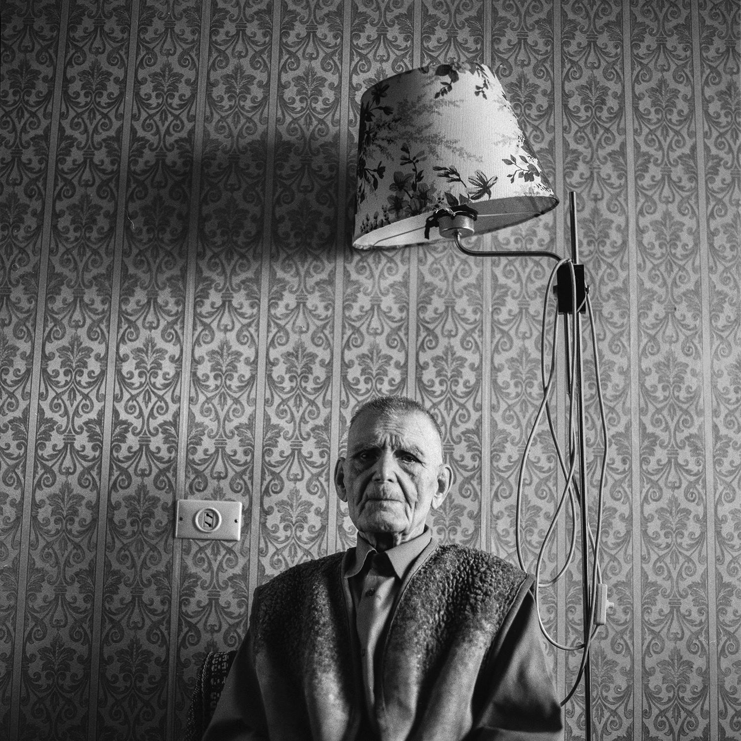 A lonely old soldier of Great Patriotic War sits in a chair under a floor lamp in warm clothes in his apartment